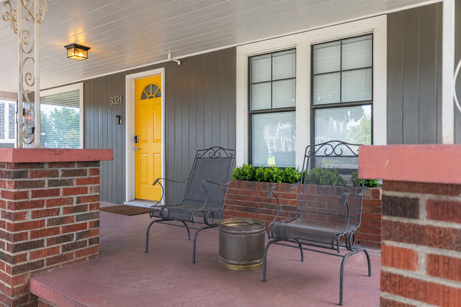 A porch with two metal chairs, a yellow door, and a brick wall.