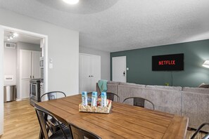 A modern living room with a wooden dining table, grey sofa, and a wall-mounted TV displaying 'NETFLIX'.