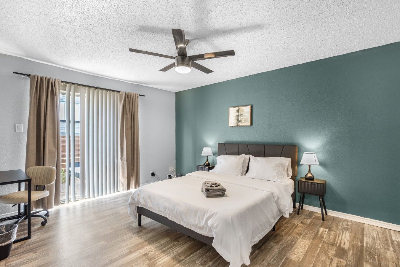 Una habitación de hotel con cama, mesitas de noche, ventilador de techo, ventana con cortinas y una silla.