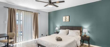 Una habitación de hotel con cama, mesitas de noche, ventilador de techo, ventana con cortinas y una silla.
