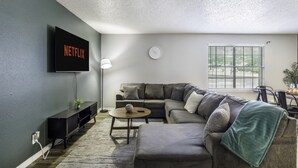 Ein Wohnzimmer mit einem Ecksofa, einem Couchtisch, einem an der Wand montierten Fernseher und einem Fenster mit Jalousien.