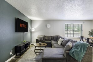 Ein Wohnzimmer mit einem Ecksofa, einem Couchtisch, einem an der Wand montierten Fernseher und einem Fenster mit Jalousien.