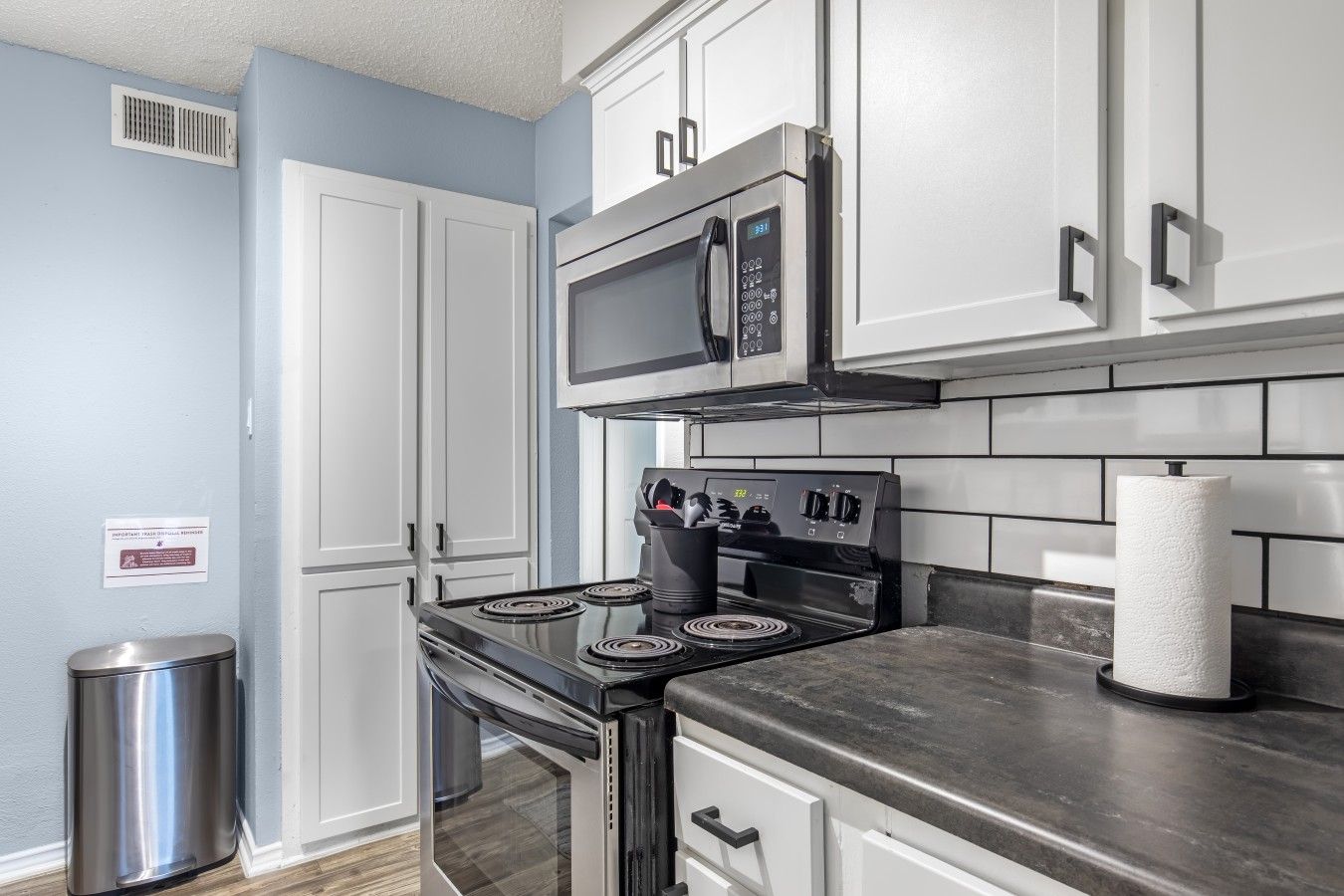 A kitchen with white cabinets, a stainless steel stove, a microwave, and a trash can.