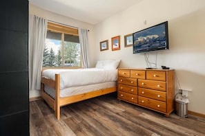 Ein Schlafzimmer mit Bett, Holzkommode, Fernseher und einem Fenster mit Vorhängen.