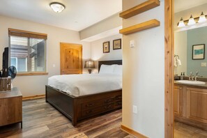 A bedroom with a bed, wooden floor, a desk, a window, and a bathroom with a sink and mirror.