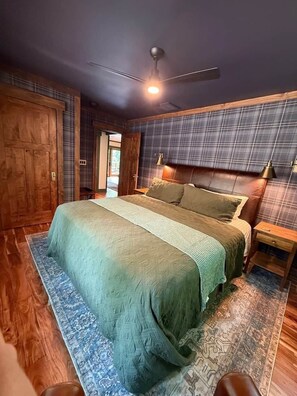 A bedroom with a large bed, wooden flooring, a ceiling fan, and a plaid-patterned wall. - Camp Shady Pines | Luxury Blue Ridge Mountain Retreat (Transylvania County)