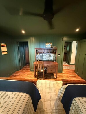 Ein Zimmer mit zwei Betten, einem Schreibtisch, einem Stuhl und einem Deckenventilator. Ein Schild mit der Aufschrift 'DO NOT PET THE FLUFFY DOGS'.