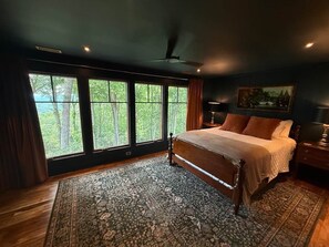 Ein Schlafzimmer mit einem großen Bett, Blick auf Bäume durch die Fenster und einem Deckenventilator.
