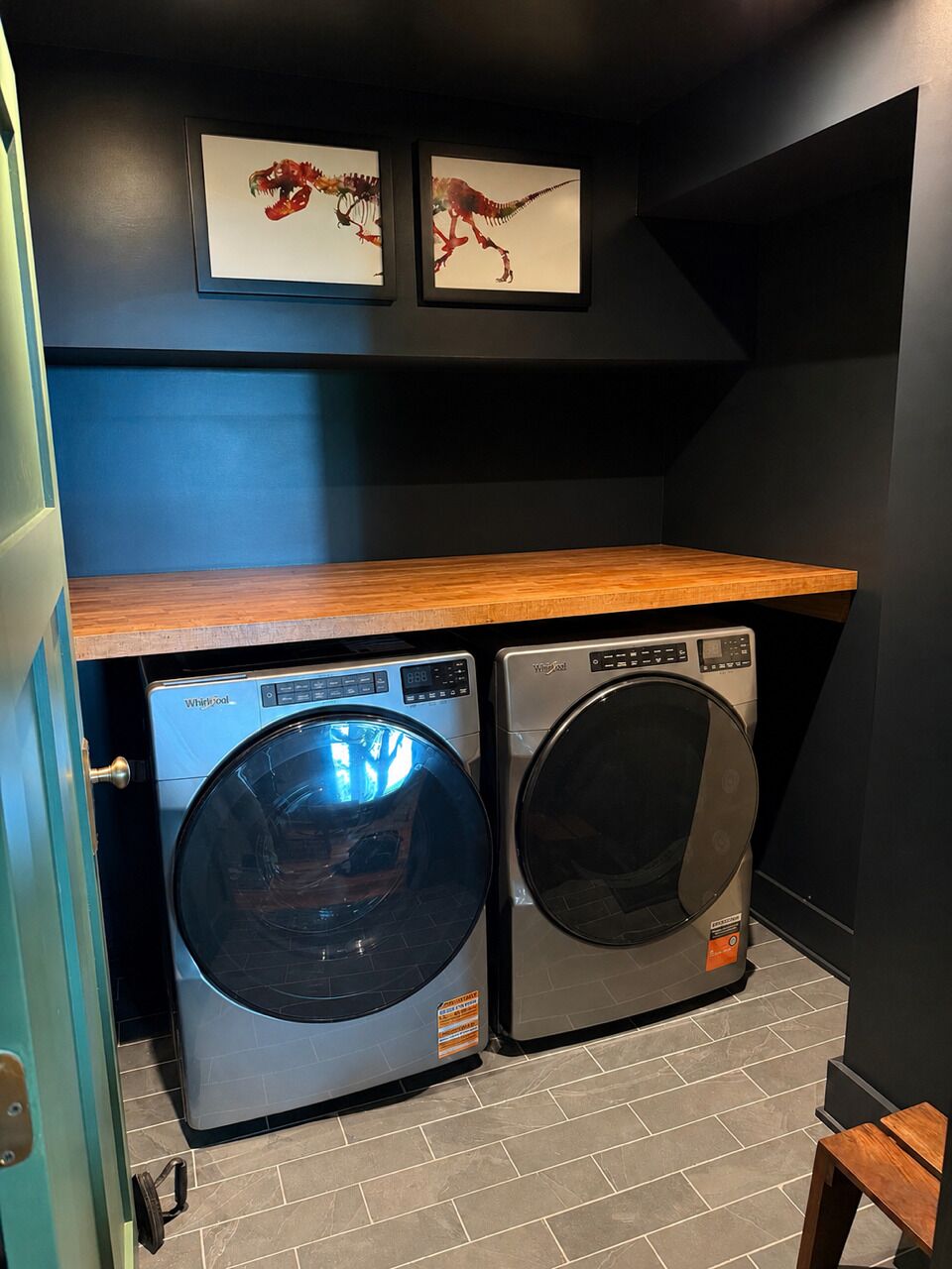 A laundry room with two Whirlpool washers, a wooden shelf above, and dinosaur-themed artwork on the wall.