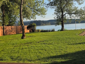 A grassy area with trees and a wooden fence, overlooking a body of water and distant hills.