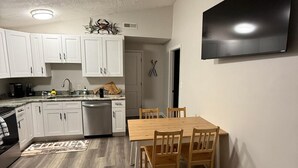 Een moderne keuken met witte kasten, een eettafel met stoelen, een flatscreen televisie aan de muur en een vloerkleed met een patroon.