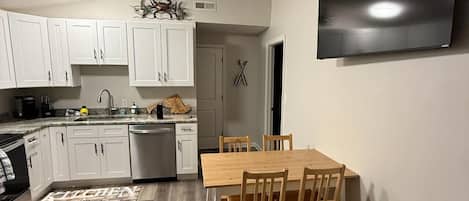 A modern kitchen with white cabinets, a dining table with chairs, a flat-screen TV mounted on the wall, and a patterned rug on the floor.