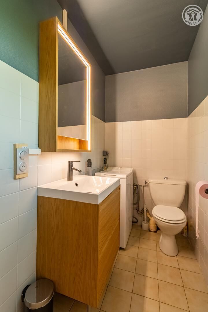 Un baño moderno con tocador de madera, un espejo grande, un lavamanos blanco, un inodoro y un pequeño cubo de basura.