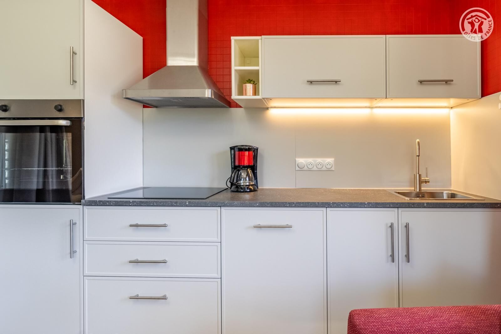 A modern kitchen with white cabinets, a stainless steel range hood, a coffee maker, and a red accent wall.