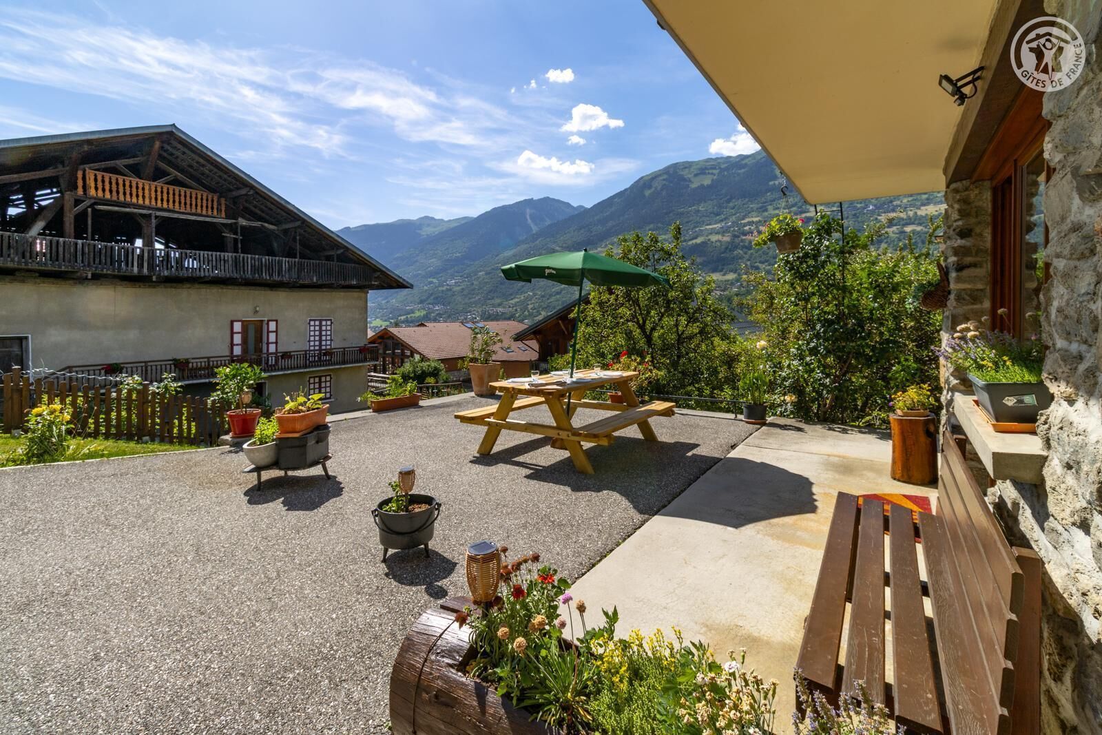 A patio with a wooden table and benches, surrounded by potted plants and a view of mountains.