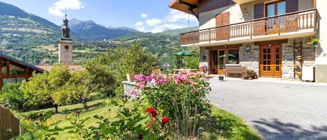 A hotel with a stone facade, wooden balcony, and a view of mountains and a church tower.