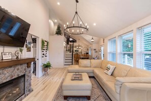 Ein Wohnzimmer mit Kamin, großem Fernseher, einer Chandelüster, einer Couch und einem Essbereich im Hintergrund.