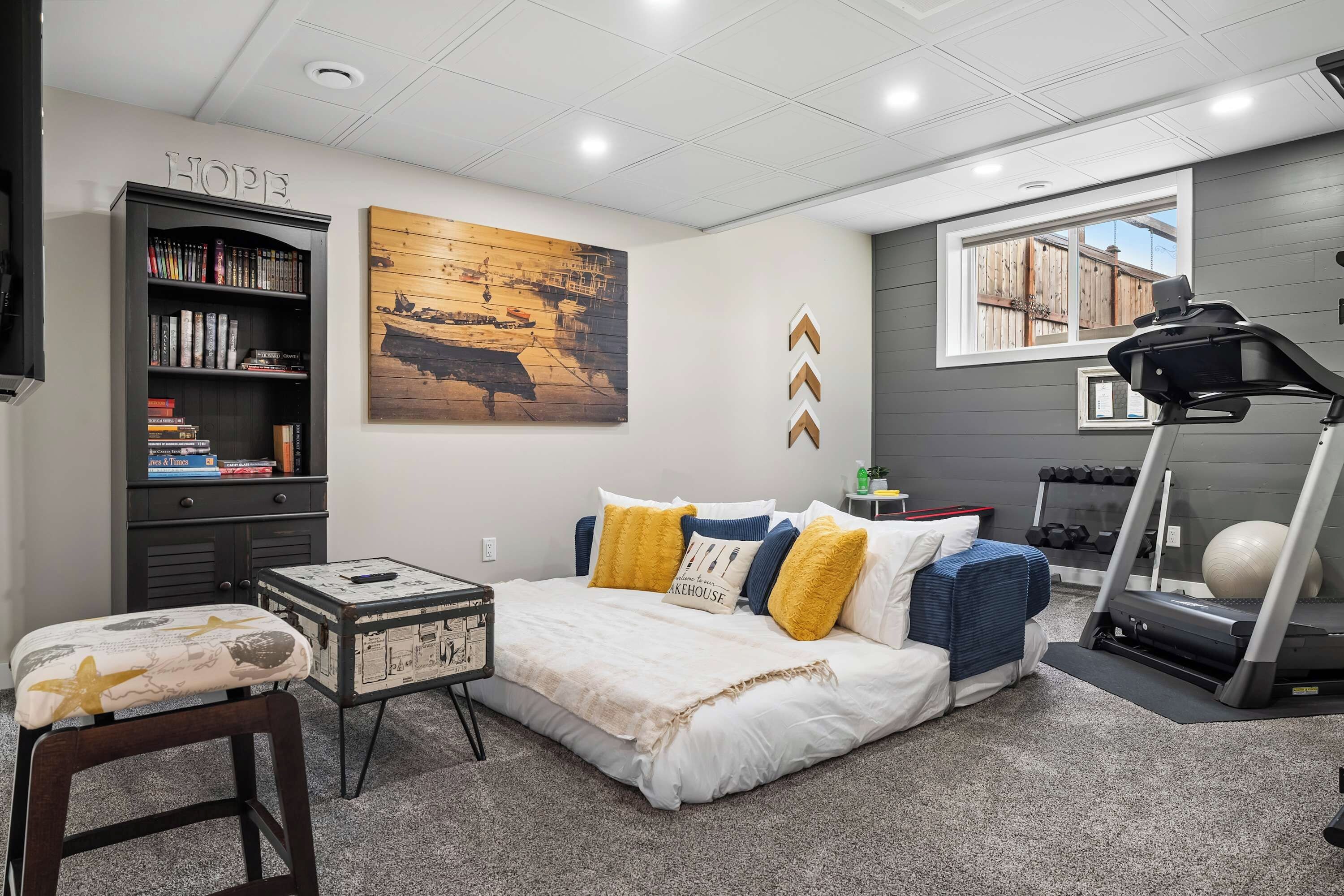 A bedroom with a bed, a bookshelf, a painting, a treadmill, and a window.