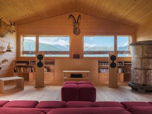 Ein Holzinnenraum mit Kamin, Bücherregalen, einem roten Hocker und Blick auf die Berge.