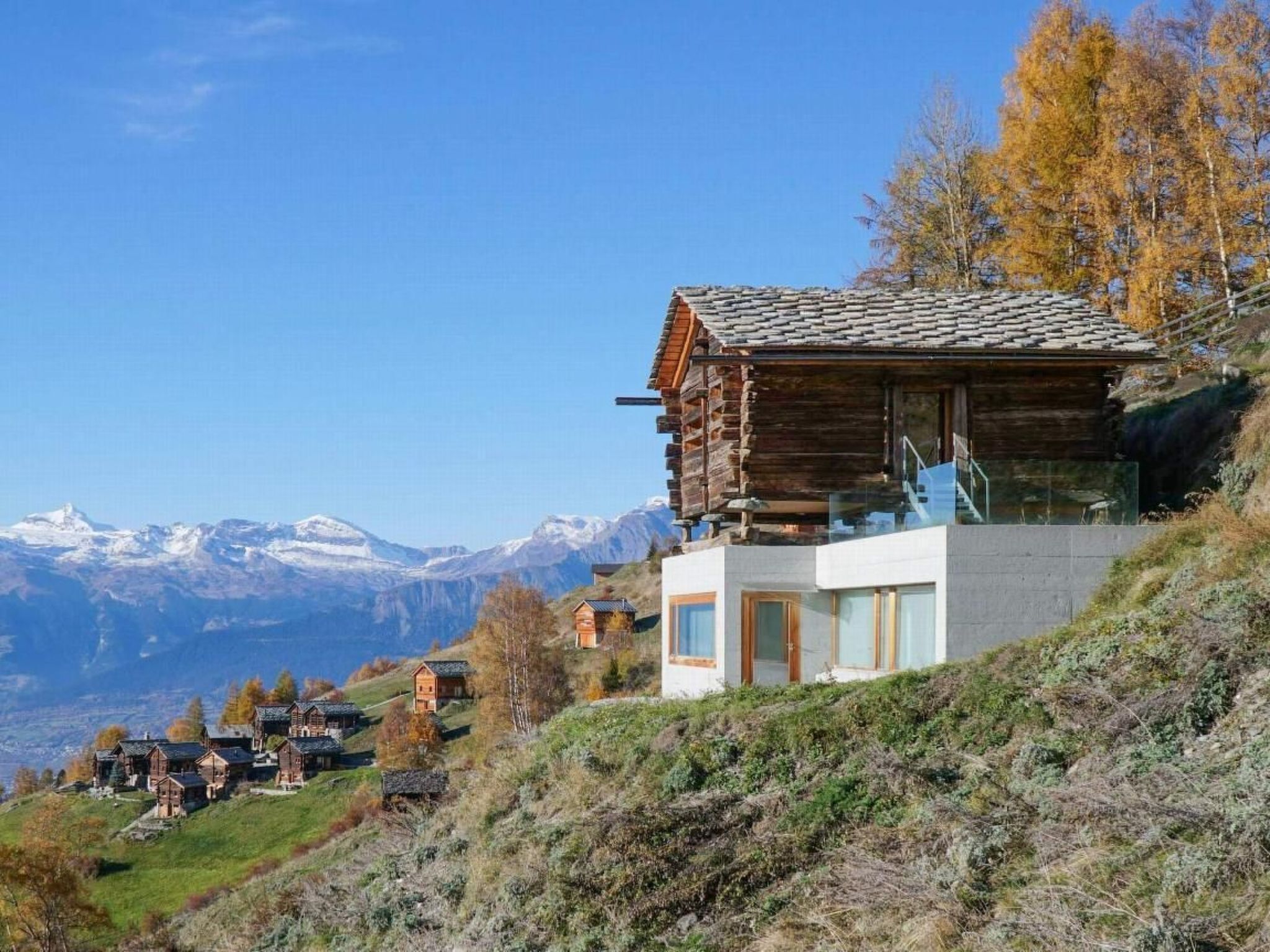 Una cabaña de madera moderna con balcón de vidrio, rodeada de un paisaje montañoso.