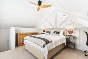 A bedroom with a sloped ceiling, a bed with a wooden headboard, bedside tables with lamps, and a chair. - The Boat House - Rustic Luxury in Matakana (Matakana)