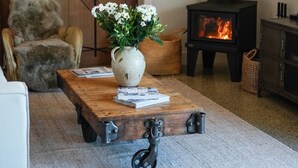 En hyggelig stue med et træbord, en hvid vase med blomster, en læderlænestol, en pejs og en stor trædør.