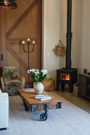 A cozy living room with a wooden coffee table, a white vase with flowers, a leather armchair, a fireplace, and a large wooden door. - The Boat House - Rustic Luxury in Matakana (Matakana)