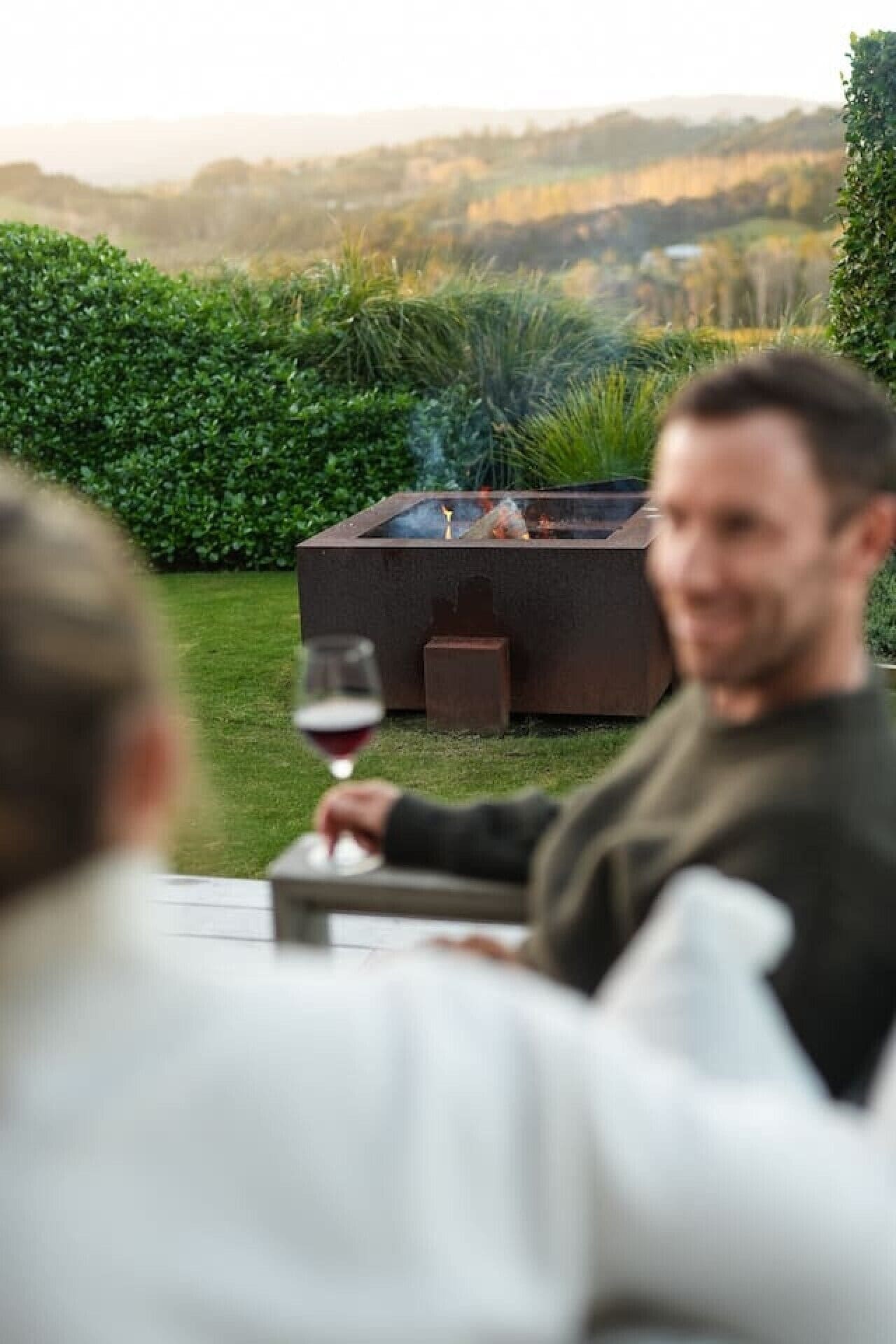 A person sitting at an outdoor table with a glass of wine, a fire pit, and a view of a hilly landscape in the background.