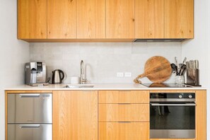 A modern kitchen with wooden cabinets, stainless steel appliances, a coffee machine, and a wooden cutting board. - The Boat House - Rustic Luxury in Matakana (Matakana)