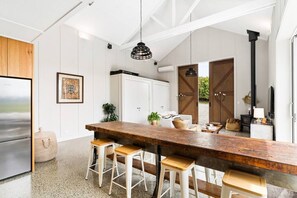 A modern kitchen with a long wooden island, white bar stools, a hanging pendant light, and a stainless steel refrigerator. - The Boat House - Rustic Luxury in Matakana (Matakana)