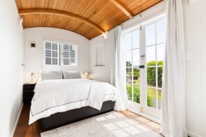 A bedroom with a bed, a nightstand, a window with shutters, and a view of greenery outside. - The Boat House - Rustic Luxury in Matakana (Matakana)