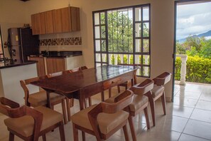 A dining area with a large wooden table and chairs, a kitchen with wooden cabinets, and a view of greenery through a window.