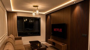 A modern living room with a sofa, a coffee table, a TV mounted on a wooden wall, and a window with blinds. - Modern 1BR Netflix • WiFi • Free Parking (Tangier)