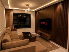 A modern living room with a sofa, a coffee table, a TV mounted on a wooden wall, and a window with blinds. - Modern 1BR Netflix • WiFi • Free Parking (Tangier)
