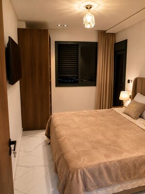A bedroom with a large bed, a TV mounted on the wall, a window with blinds, and a nightstand with a lamp. - Modern 1BR Netflix • WiFi • Free Parking (Tangier)