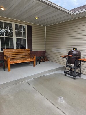 A covered patio area with a wooden bench, a grill cart, and a stack of firewood.