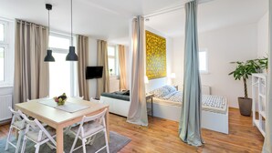 A modern room with a wooden floor, a dining table with chairs, a bed with a headboard, a TV, and a large window with curtains.