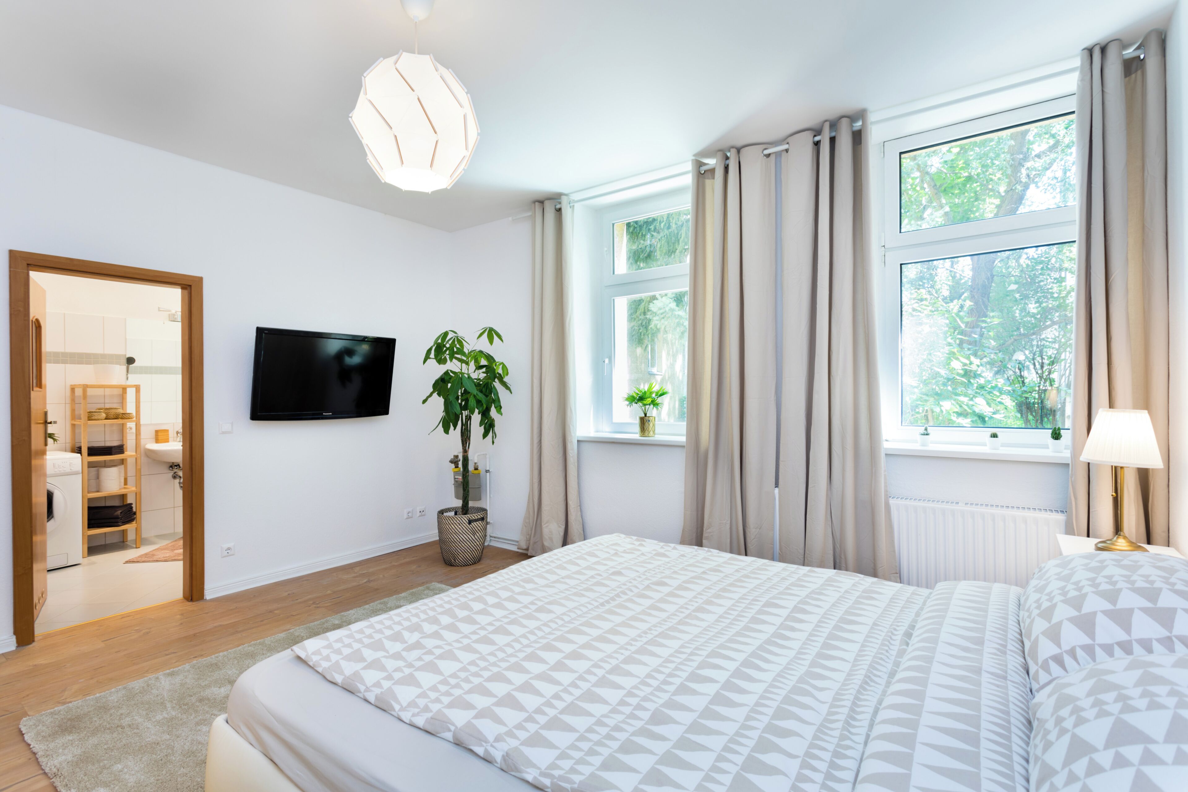 A modern bedroom with a bed, a TV, a potted plant, and a window with curtains.