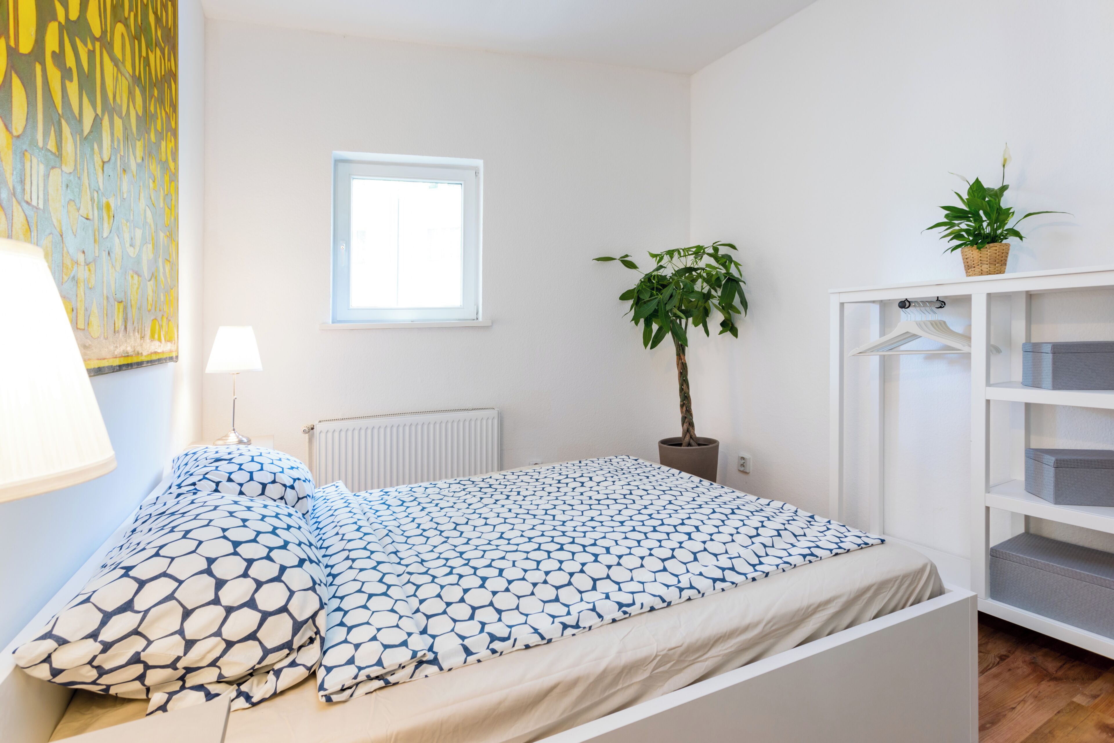 A bedroom with a bed, a radiator, a window, a wall art piece, a potted plant, and a shelving unit.