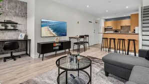 Een moderne woonkamer met een televisie, een ronde salontafel, een grijze bank en een keukenhoek met houten kasten en barstoelen.
