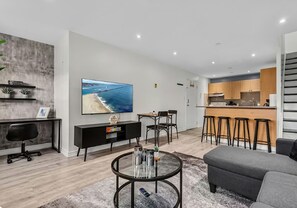 A modern living room with a TV, a round coffee table, a grey sofa, and a kitchen area with wooden cabinets and bar stools. - 2BR Home | Free Parking | Fast WiFi (Toronto)