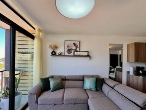 Ein modernes Wohnzimmer mit einem Ecksofa, einer Wandregal mit Dekoration und einem großen Fenster mit Blick ins Freie.