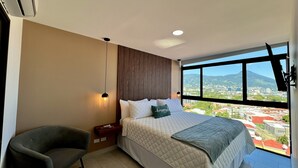 Una camera da letto moderna con un letto grande, una poltrona, un comodino e vista sulla città attraverso un'ampia finestra.
