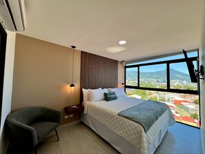 Ein modernes Schlafzimmer mit einem großen Bett, einem Sessel, einem Nachttisch und Blick auf eine Skyline durch ein großes Fenster.
