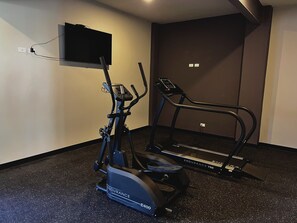 A home gym with an elliptical and a treadmill, both with control panels, against a plain wall with a mounted TV. - Cozy 1-bedroom apartment with AC in fantastic San Salvador (San Salvador)