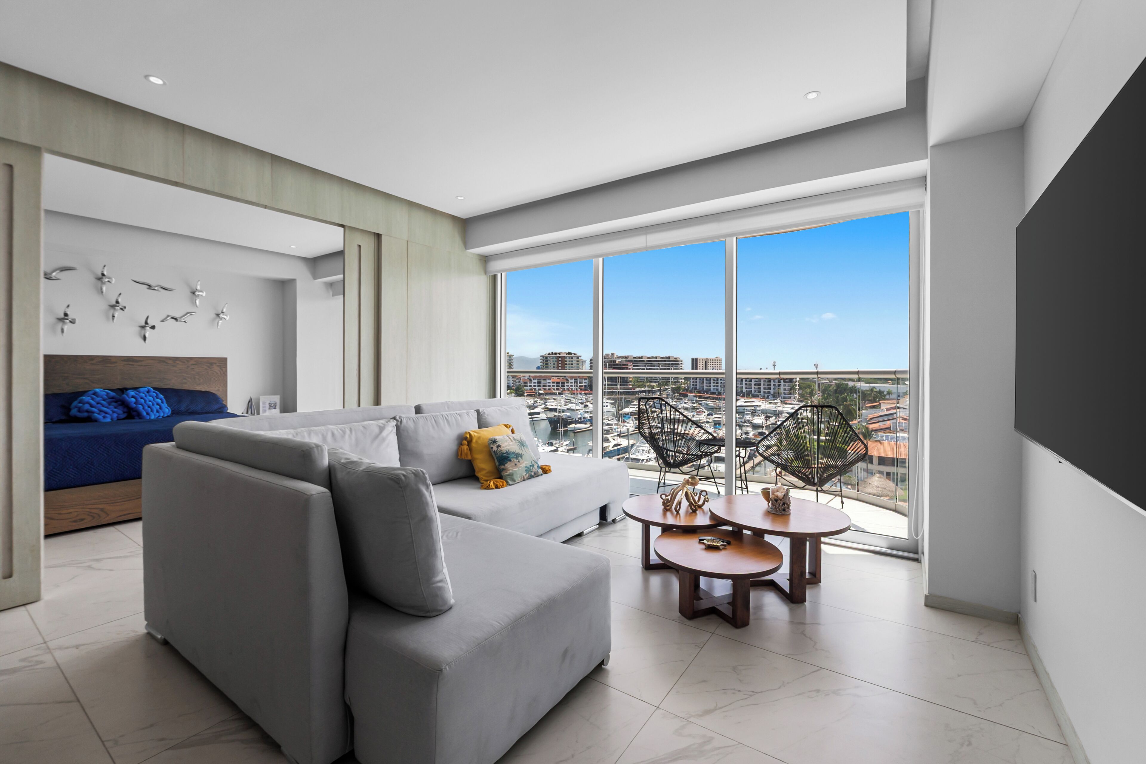 A modern living room with a grey sofa, a coffee table, and a view of a marina through large windows.
