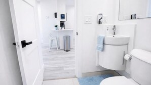 A modern bathroom with a toilet, sink, and mirror. There is a trash bin and a small table with a chair in the background. - Wavecrest Retreat On Strip  (Daytona Beach)