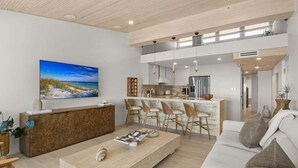 A modern living room with a wooden reception desk, a large flat-screen TV, a sofa, and a kitchen area with bar stools. - Bay Pearl - Bradenton Beach Condo by Anna Maria Life Vacation Rentals (Bradenton Beach)