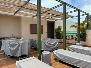 Uma área externa com mesas e cadeiras cobertas, estrutura de pérgola e vista dos edifícios ao redor.