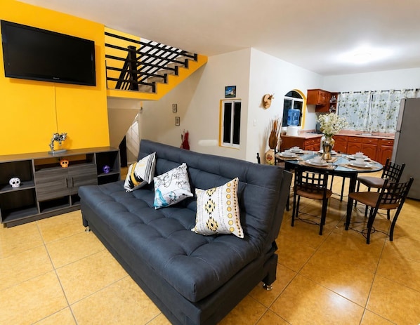A living room with a sofa, TV, staircase, dining table, chairs, and kitchen area.